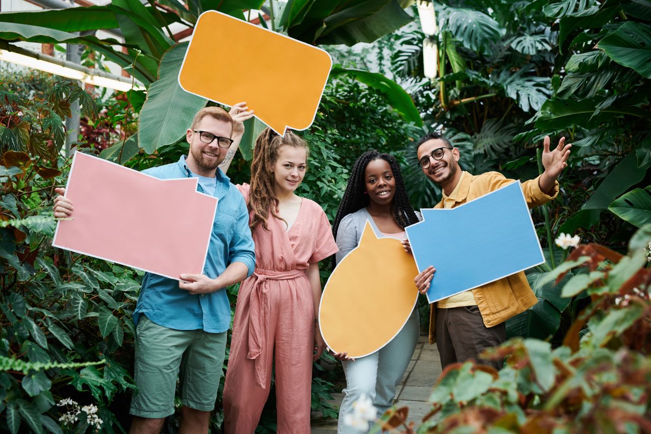 A diverse group of friends holding colorful speech bubble signs in a lush greenhouse.